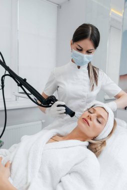Genç bir kız güzellik salonunda yüzünün derisi için kömür soyuyordu. Lazer darbeleri deriyi temizler. Donanım kozmetolojisi. Fototermoliz süreci, deriyi ısıtır..