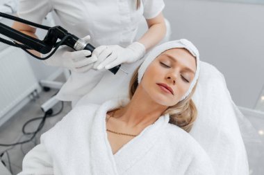 Genç bir kız güzellik salonunda yüzünün derisi için kömür soyuyordu. Lazer darbeleri deriyi temizler. Donanım kozmetolojisi. Fototermoliz süreci, deriyi ısıtır..