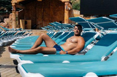 Genç bir atletik adam mutlu bir şekilde gülümsüyor ve güneşli bir otelde güneşli bir günde güneşleniyor. Oteldeki aile tatiliniz kutlu olsun. Yaz tatilleri ve turizm.