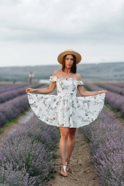 Narin elbiseli ve şapkalı genç bir kız güzel bir lavanta tarlasında yürüyor ve çiçeklerin kokusundan hoşlanıyor. Dinlenme ve güzel doğa. Lavanta açması ve çiçek toplaması..