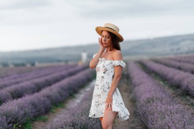 Narin elbiseli ve şapkalı genç bir kız güzel bir lavanta tarlasında yürüyor ve çiçeklerin kokusundan hoşlanıyor. Dinlenme ve güzel doğa. Lavanta açması ve çiçek toplaması..