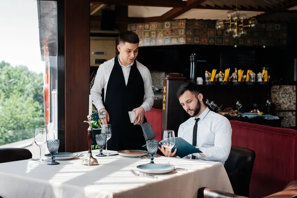 高級レストランの若いビジネスマンがメニューを調べ スタイリッシュなエプロンで若いウェイターに注文します カスタマーサービス レストランでのテーブルサービス — ストック写真