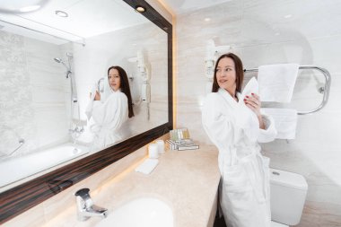 Genç ve güzel bir kız yüzünü yıkar ve güzel beyaz bir banyoda havlu tutar. Otelde taze bir günaydın. Dinlen ve seyahat et. Otel eğlencesi ve turizm..