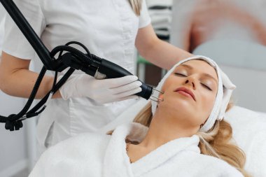 Genç bir kız güzellik salonunda yüzünün derisi için kömür soyuyordu. Lazer darbeleri deriyi temizler. Donanım kozmetolojisi. Fototermoliz süreci, deriyi ısıtır..
