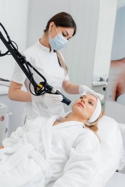 Genç bir kız güzellik salonunda yüzünün derisi için kömür soyuyordu. Lazer darbeleri deriyi temizler. Donanım kozmetolojisi. Fototermoliz süreci, deriyi ısıtır..
