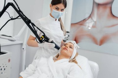 Genç bir kız güzellik salonunda yüzünün derisi için kömür soyuyordu. Lazer darbeleri deriyi temizler. Donanım kozmetolojisi. Fototermoliz süreci, deriyi ısıtır..