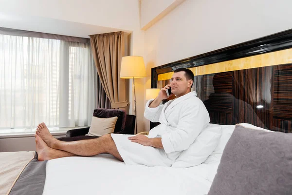 A young guy in a white lab coat is sitting on the bed and talking on the phone in his hotel room. Rest and travel. Hotel rest, and spa treatments.