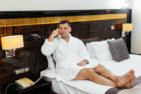 Beyaz laboratuvar önlüklü genç bir adam otel odasında yatağında oturmuş telefonla konuşuyor. Dinlen ve seyahat et. Otel istirahatı ve spa tedavisi..