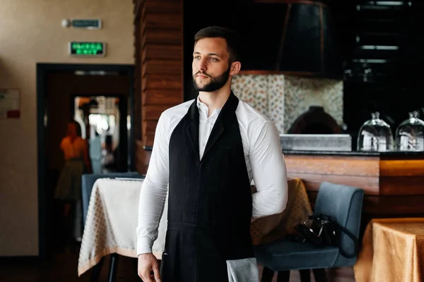 A young waiter with a beard puts on an apron and prepares for a working day in a fine restaurant.