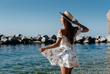 Şapkalı ve sırtında açık renk elbisesi olan güzel bir kadın güneşli bir günde okyanus kıyısında büyük kayaların arkasında yürüyor. Turizm ve tatil gezisi.