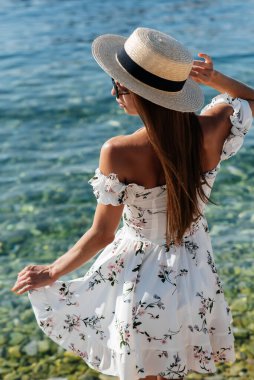 Şapkalı ve sırtında açık renk elbisesi olan güzel bir kadın güneşli bir günde okyanus kıyısında büyük kayaların arkasında yürüyor. Turizm ve tatil gezisi.