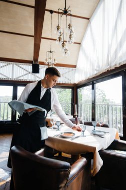 Şık üniformalı genç bir erkek garson güzel bir gurme restoranında masaya servis yapmakla meşgul. Restoran faaliyetleri, en üst düzeyde..