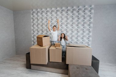 Erkek ve kız kardeş kanepedeki karton kutuların arka planına karşı yeni bir daireye taşınıp eğleniyorlar. Gayrimenkul alımı. Ev partisi, teslimat..