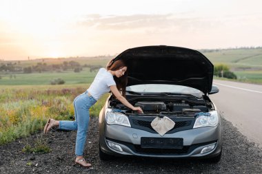 A young girl stand near a broken car in the middle of the highway during sunset and tries to repair it. Troubleshooting the problem. Car service. Car breakdown on the road. clipart