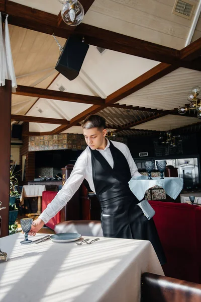 Şık üniformalı genç bir erkek garson güzel bir gurme restoranında masaya servis yapmakla meşgul. Restoran faaliyetleri, en üst düzeyde..