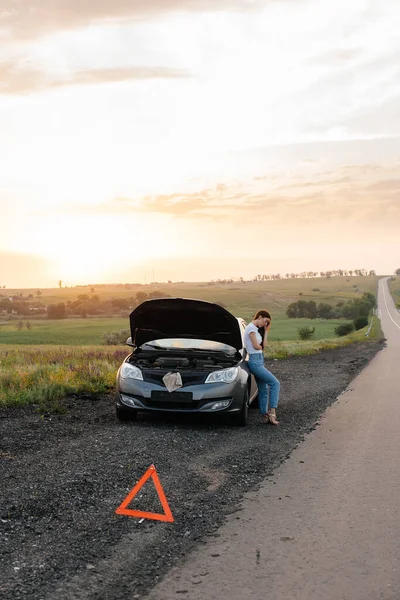 A frustrated young girl stands near a broken-down car in the middle of the highway during sunset. Breakdown and repair of the car. Waiting for help. Car service. Car breakdown on road.