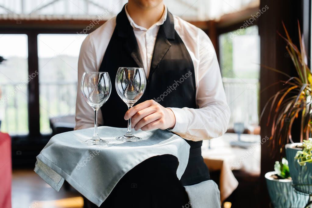 A young waiter in a stylish uniform stands with glasses on a tray near the table in a beautiful gourmet restaurant close-up. Restaurant activity, of the highest level.