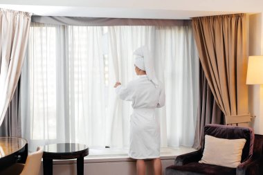 Beyaz önlüklü güzel bir kız otel odasının penceresinin yanında duruyor ve kahve içiyor. Dinlen ve seyahat et. Otel eğlencesi ve turizm..