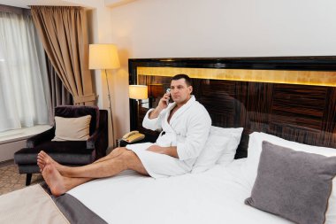 Beyaz laboratuvar önlüklü genç bir adam otel odasında yatağında oturmuş telefonla konuşuyor. Dinlen ve seyahat et. Otel istirahatı ve spa tedavisi..