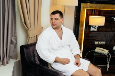 Beyaz önlüklü genç bir adam otel odasının penceresinin yanında oturuyor. Dinlen ve seyahat et. Otel eğlencesi ve turizm..