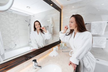 Genç güzel bir kız güzel beyaz bir banyoda dişlerini fırçalıyor. Otelde taze bir günaydın. Dinlen ve seyahat et. Otel eğlencesi ve turizm..