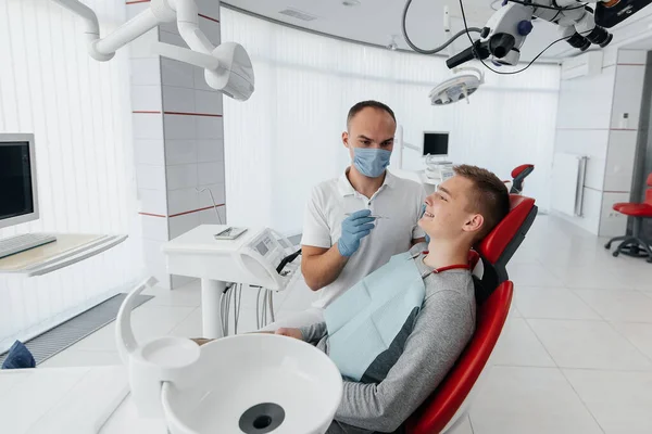 若い歯科医は 近代的な白い歯科のクローズアップで若い男の歯を調べ 扱います 歯科用義歯 治療と歯のホワイトニング 現代の歯科医 — ストック写真