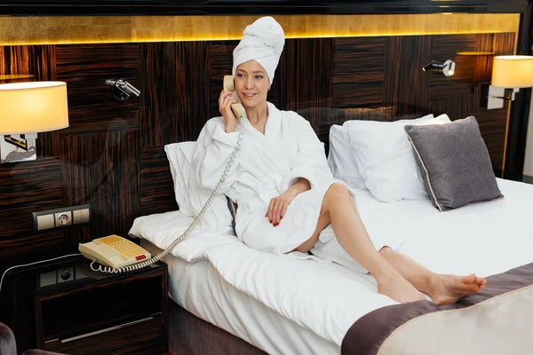 A young beautiful girl in a white coat is talking on the phone in her hotel room. Rest and travel. Hotel recreation, and tourism.