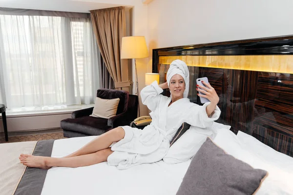 A young beautiful girl in a white coat is talking on the phone in her hotel room. Rest and travel. Hotel recreation, and tourism.