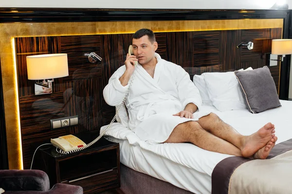 A young guy in a white lab coat is sitting on the bed and talking on the phone in his hotel room. Rest and travel. Hotel rest, and spa treatments.