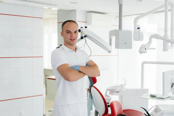 若い歯科医は赤い歯の椅子の近くに立っており 現代の白い歯科で笑顔 若者からの介護の治療と予防 現代の歯科と義肢装具 — ストック写真