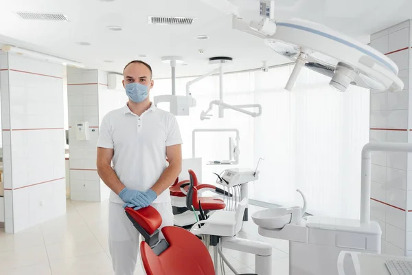 マスクの若い歯科医は 赤い歯の椅子の近くに立っており 現代の白い歯科で笑顔 現代の歯科と義肢装具 — ストック写真
