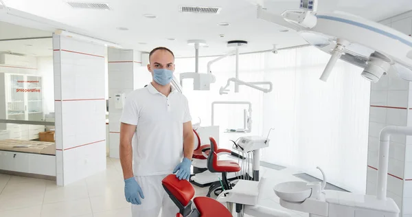 マスクの若い歯科医は 赤い歯の椅子の近くに立っており 現代の白い歯科で笑顔 現代の歯科と義肢装具 — ストック写真