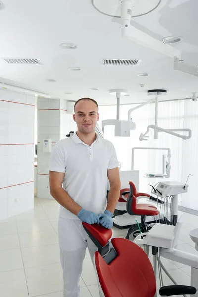 若い歯科医は赤い歯の椅子の近くに立っており 現代の白い歯科で笑顔 若者からの介護の治療と予防 現代の歯科と義肢装具 — ストック写真