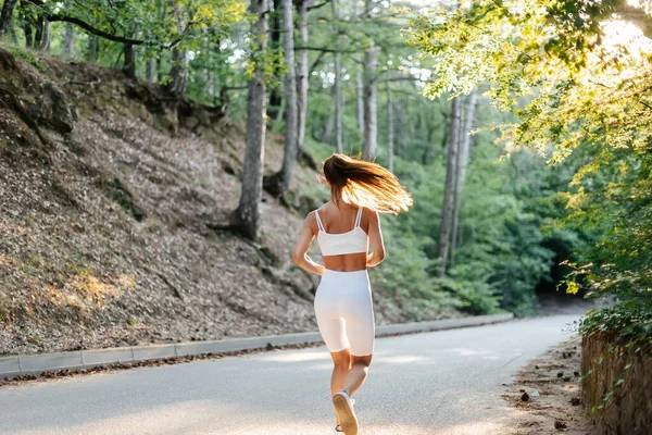 白いスポーツ服を着た若い美しい少女が 日没の間 鬱蒼とした森の中の道路で彼女と一緒に走っています 新鮮な空気の中でスポーツをしている 健康的なライフスタイル — ストック写真