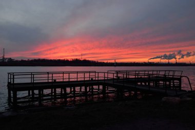 Gün batımında nehir. Dramatik gökyüzü, parlayan kırmızı bulutlar. Arka planda port terminali. Kış manzarası kavramı. Doğa, ekoloji, çevresel hasar, kirlilik, iklim değişikliği