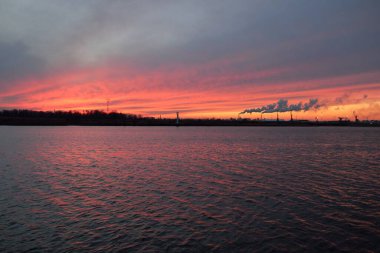 Gün batımında nehir. Dramatik gökyüzü, parlayan kırmızı bulutlar. Arka planda port terminali. Kış manzarası kavramı. Doğa, ekoloji, çevresel hasar, kirlilik, iklim değişikliği