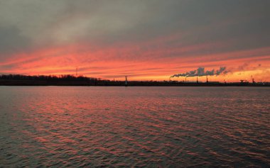 Gün batımında nehir. Dramatik gökyüzü, parlayan kırmızı bulutlar. Arka planda port terminali. Kış manzarası kavramı. Doğa, ekoloji, çevresel hasar, kirlilik, iklim değişikliği