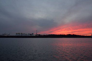 Gün batımında nehir. Dramatik gökyüzü, parlayan kırmızı bulutlar. Arka planda port terminali. Kış manzarası kavramı. Doğa, ekoloji, çevresel hasar, kirlilik, iklim değişikliği