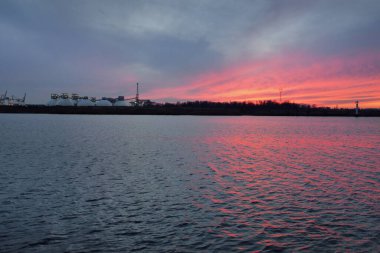 Gün batımında nehir. Dramatik gökyüzü, parlayan kırmızı bulutlar. Arka planda port terminali. Kış manzarası kavramı. Doğa, ekoloji, çevresel hasar, kirlilik, iklim değişikliği