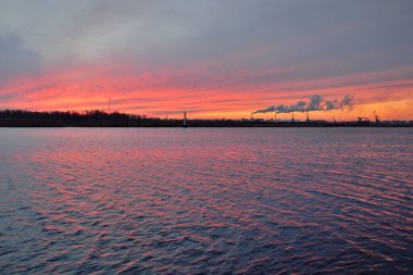 Gün batımında nehir. Dramatik gökyüzü, parlayan kırmızı bulutlar. Arka planda port terminali. Kış manzarası kavramı. Doğa, ekoloji, çevresel hasar, kirlilik, iklim değişikliği