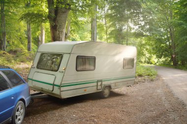 Beyaz karavan karavanı ve muhteşem ağaçların altındaki kırsal yolda park edilmiş bir araba. Orman kamp alanı. Avrupa. Yaşam tarzı, özgürlük, seyahat, eko-turizm, yolculuk, dinlenme, ulaşım, karavan, karavan