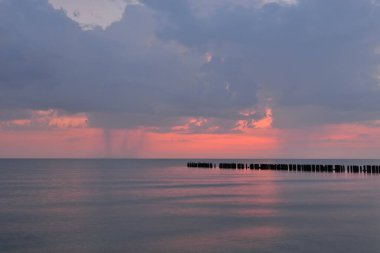 Fırtınadan sonra Baltık kıyılarından panoramik manzara. Dramatik gökyüzü, parlayan pembe günbatımı bulutları. Eski ahşap iskele. Doğa, çevre, ekoloji, iklim değişikliği, kararsız hava