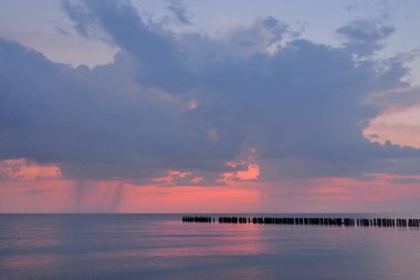Fırtınadan sonra Baltık kıyılarından panoramik manzara. Dramatik gökyüzü, parlayan pembe günbatımı bulutları. Eski ahşap iskele. Doğa, çevre, ekoloji, iklim değişikliği, kararsız hava