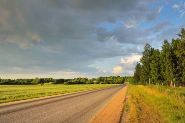 Gün batımında tarım arazisi ve ormanın içinden geçen şehir otoyolu. Fırtınadan sonra dramatik bir gökyüzü. Sonbaharın başında. Yumuşak akşam güneşi. Tatiller, yol gezisi, sürüş, yolculuk