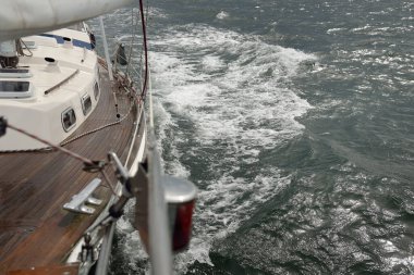 Fırtınada açık denizde yelken açan eski klasik bir yat. Tahta meşe güvertesi. Direğe ve yelkenlere bakın. Ketçap yelkenli. Dalgalar, su sıçramaları. Gezi, tatil, yelkenli yarışı, spor, boş zaman aktivitesi.