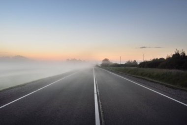 Boş bir otoyol (yeni asfalt yol) gün batımında sisli bir havada tarlalar ve ormanlar arasında. Yumuşak güneş ışığı. Kırsal alan. Uzak yerler, köy, yol gezisi, turizm, rota, lojistik, özgürlük kavramları