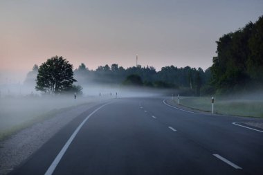 Gün batımında sisli bir yolda (asfalt yol). Ayın doğuşu, alacakaranlık, dramatik gökyüzü. Idyllic yaz kırsal manzarası. Ulaşım, uzak yerler, tehlikeli sürüş