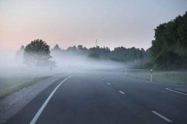 Gün batımında sisli bir yolda (asfalt yol). Ayın doğuşu, alacakaranlık, dramatik gökyüzü. Idyllic yaz kırsal manzarası. Ulaşım, uzak yerler, tehlikeli sürüş