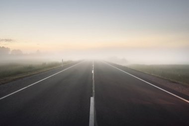 Boş bir otoyol (yeni asfalt yol) gün batımında sisli bir havada tarlalar ve ormanlar arasında. Yumuşak güneş ışığı. Kırsal alan. Uzak yerler, köy, yol gezisi, turizm, rota, lojistik, özgürlük kavramları