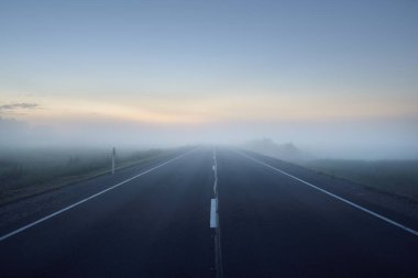 Gün batımında sisli bir yolda (asfalt yol). Ayın doğuşu, alacakaranlık, dramatik gökyüzü. Idyllic yaz kırsal manzarası. Ulaşım, uzak yerler, tehlikeli sürüş